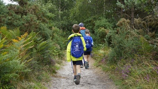 Two boys following eachother through the woodland at Dunwich Heath. The one closest to the camera is wearing a lime green anorak and carrying a blue tracker pack in a rucksack on his back.